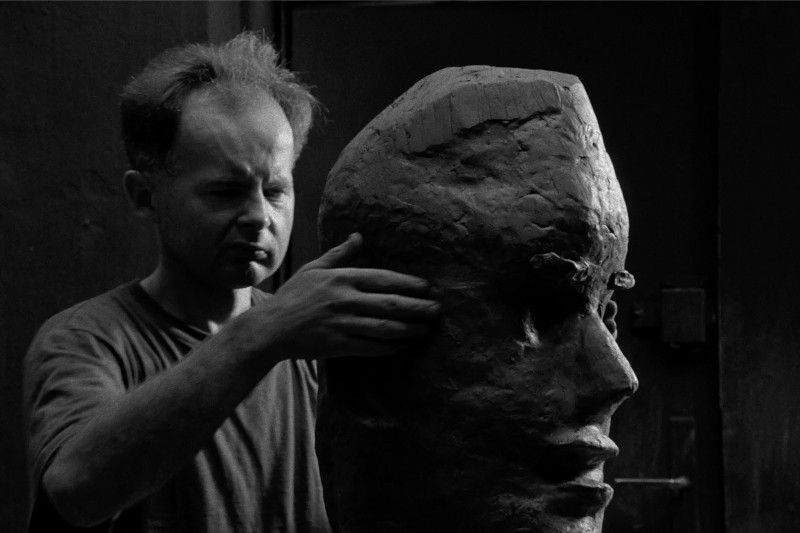 Mężczyzna z zamkniętymi oczami ostrożnie rzeźbi dużą glinianą głowę, dotykając jej twarzy jedną ręką, w słabo oświetlonym pomieszczeniu.