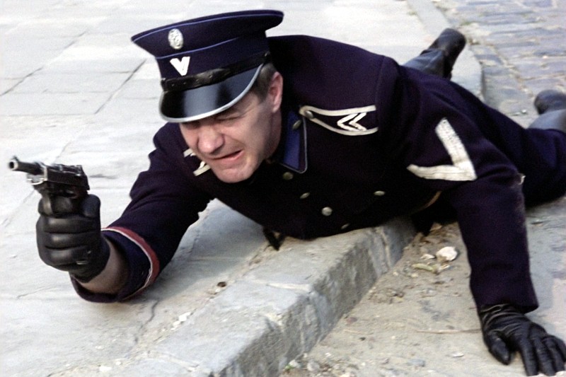 Mężczyzna w ciemnym mundurze policyjnym leży na ziemi, celując z pistoletu i sięgając ponad krawężnik. Wydaje się spięty i skupiony, ma na sobie czapkę policyjną i rękawiczki na miejskiej ulicy.