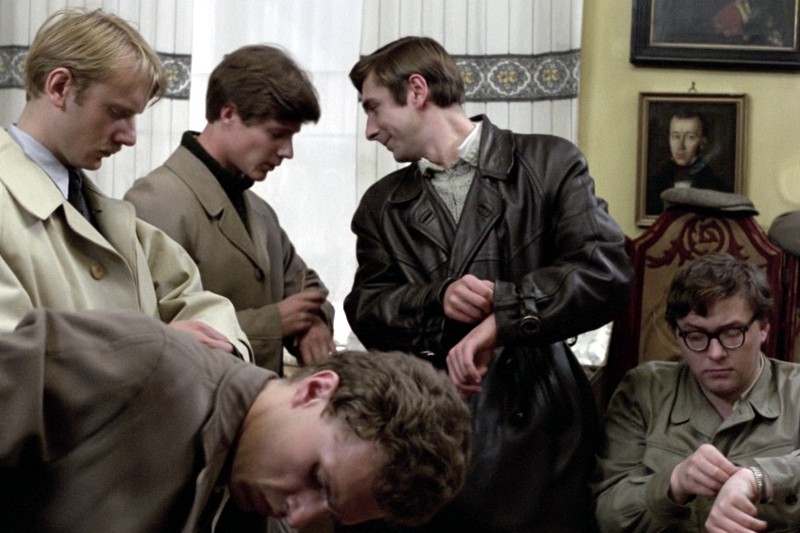 Pięciu mężczyzn ubranych w trencze i kurtki stoi i siedzi w pokoju, wszyscy patrzą na swoje nadgarstki, jakby sprawdzali godzinę. W tle widać zasłonięte okno i oprawione zdjęcia na żółtej ścianie.