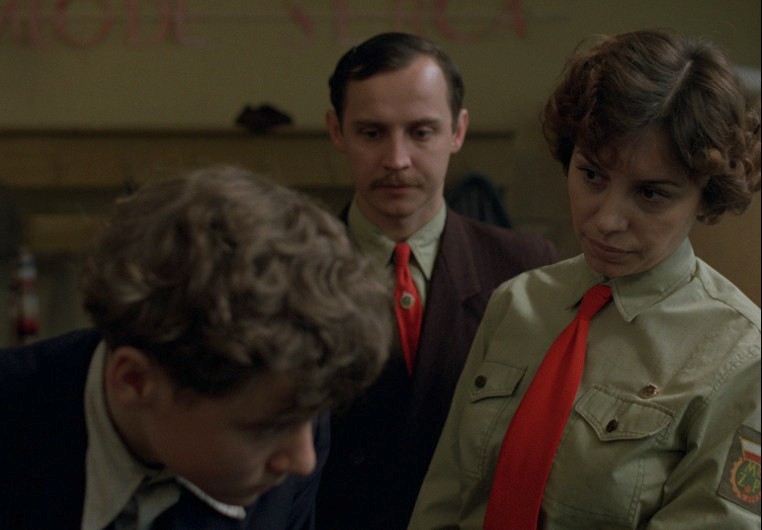 Kobieta w zielonej koszuli, mundurze i czerwonym krawacie stoi z poważnym wyrazem twarzy obok chłopca, który patrzy w dół, podczas gdy mężczyzna w garniturze i czerwonym krawacie obserwuje to w tle.