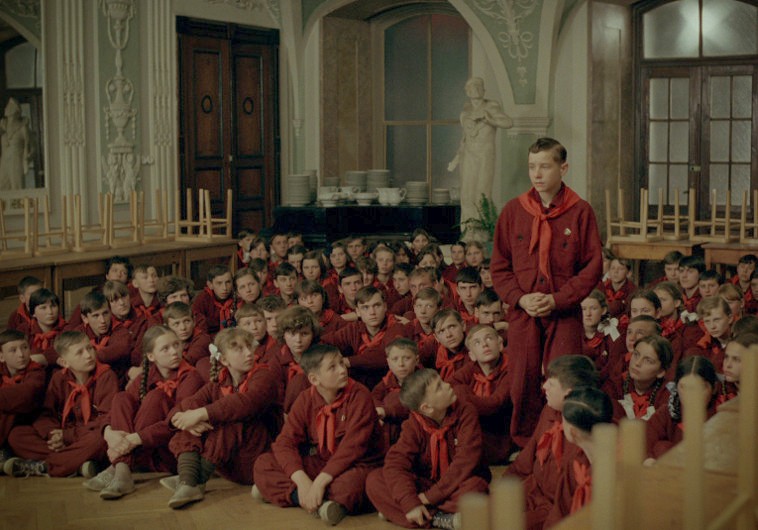Grupa dzieci ubranych w pasujące do siebie bordowe mundurki z czerwonymi szalikami siedzi na podłodze ozdobnego pokoju. Jeden chłopiec stoi na środku, jakby zwracał się do grupy. W pokoju znajdują się posągi, wysokie okna i drewniane krzesła.