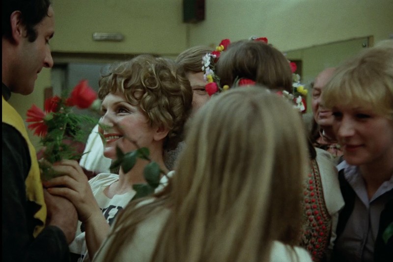 Uśmiechnięta kobieta trzymająca czerwone kwiaty jest otoczona ludźmi, niektórzy z nich mają na sobie wianki z kwiatów, a całość rozgrywa się w ciepłym, oświetlonym pomieszczeniu, co sugeruje uroczystość lub spotkanie.