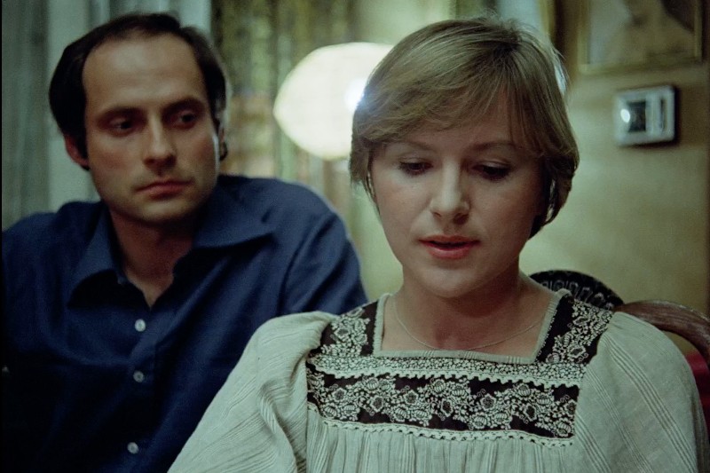 Kobieta z krótkimi blond włosami i białą haftowaną bluzką siedzi i patrzy w dół, podczas gdy mężczyzna w ciemnoniebieskiej koszuli siedzi za nią i uważnie ją obserwuje. Sceneria wydaje się być wewnątrz, z miękkim oświetleniem.