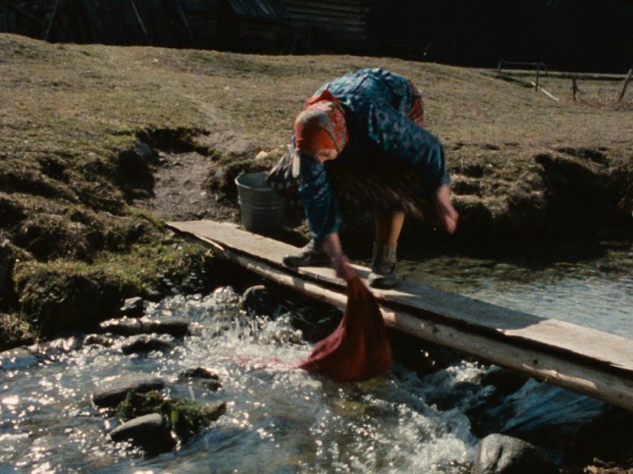 Starsza kobieta w tradycyjnym stroju pochyla się nad małym strumieniem, piorąc czerwony materiał. Stoi na drewnianym moście z metalowym wiadrem w pobliżu, otoczona trawiastym terenem.