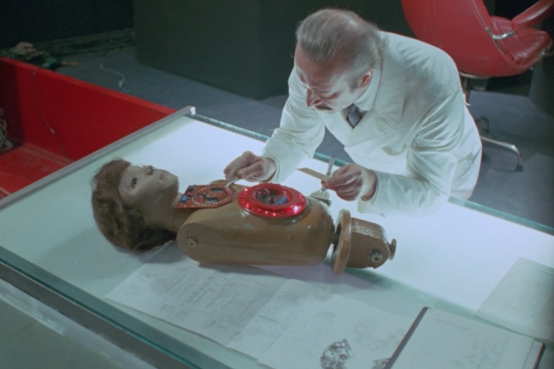 Mężczyzna w białym fartuchu laboratoryjnym bada otwartą klatkę piersiową humanoidalnego robota leżącego na szklanym stole. Wewnątrz robota widoczne są przewody i części mechaniczne.