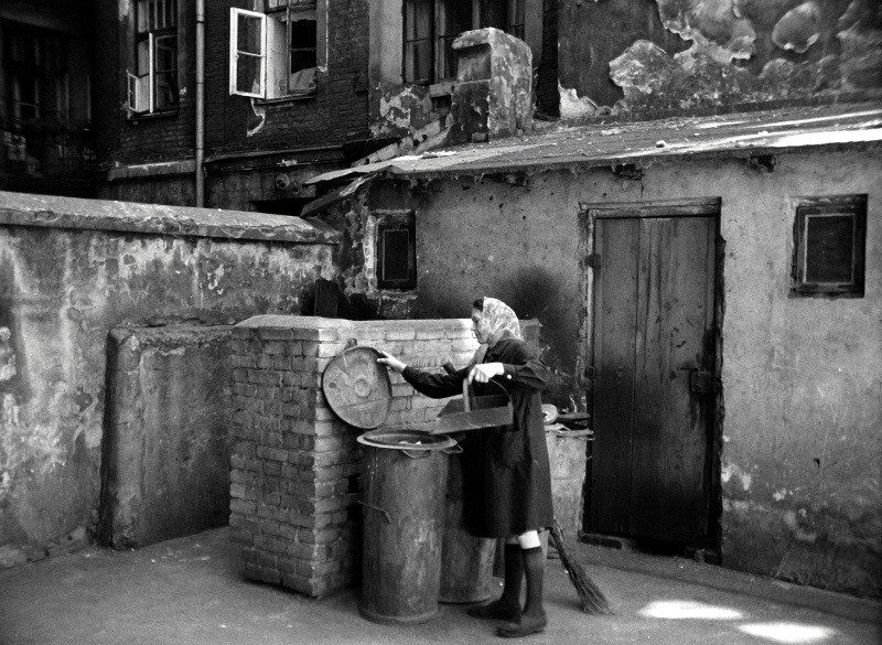 Starsza kobieta w chuście na głowie wyrzuca śmieci do metalowego pojemnika na starym, zaniedbanym podwórku z popękanymi ścianami, zabitymi deskami oknami i łuszczącą się farbą.