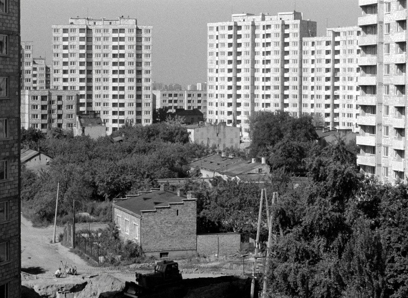 Czarno-białe zdjęcie bloków mieszkalnych z czasów sowieckich otaczających skupisko drzew i niewielkich, starszych budynków. Na pierwszym planie widać drogę gruntową i sprzęt budowlany.