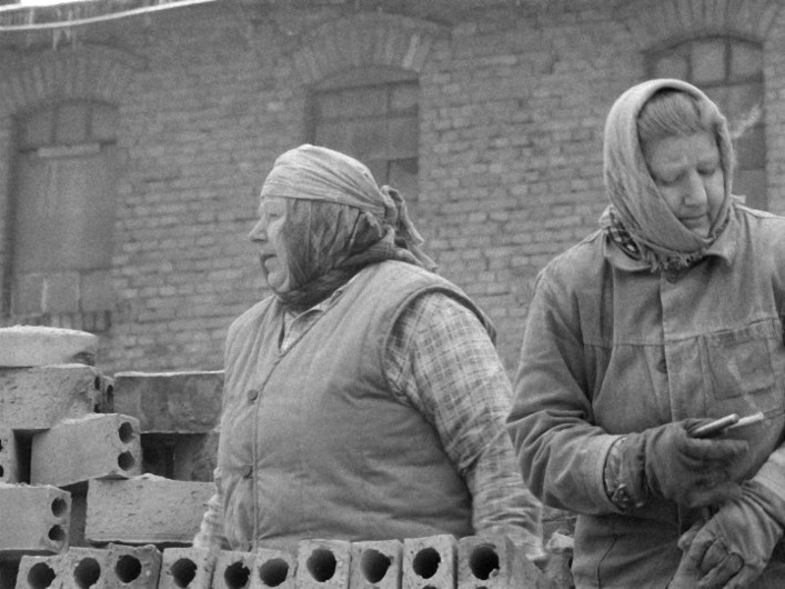 Dwie kobiety w chustach na głowach i ciepłych ubraniach stoją obok stosu cegieł przed ceglanym budynkiem; jedna patrzy w bok, druga trzyma mały przedmiot, prawdopodobnie narzędzie lub telefon.