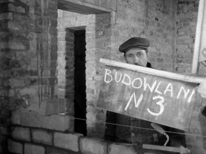 Mężczyzna w czapce z daszkiem stoi za częściowo wybudowaną ścianą z cegły. Na ręcznie napisanym znaku po polsku widnieje napis „BUDOWLANI N3”. Sceneria wydaje się być placem budowy z odsłoniętymi ścianami z cegły.