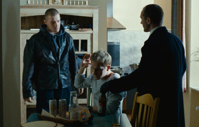 Trzech mężczyzn jest w kuchni. Jeden stoi w skórzanej kurtce, drugi siedzi przy stole z puszkami po piwie i przekąskami, podnosząc rękę, podczas gdy trzeci, w ciemnym płaszczu, stoi obok niego. Nastrój wydaje się napięty.