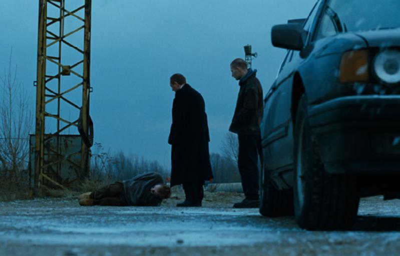 Dwóch mężczyzn w ciemnych płaszczach stoi nad osobą leżącą na ziemi w pobliżu zardzewiałej metalowej konstrukcji, a w pobliżu stoi ciemny samochód w ponury, pochmurny dzień. Scena wydaje się napięta i dramatyczna.