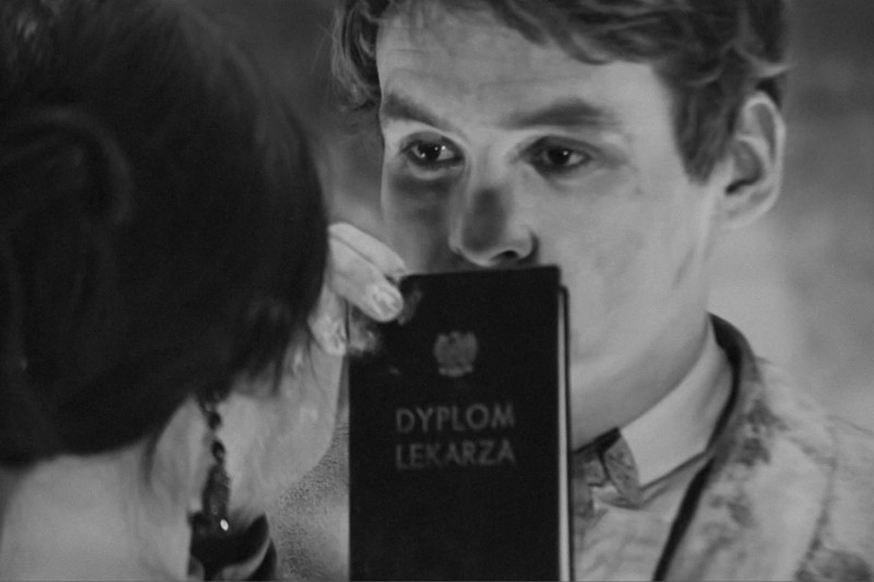 Mężczyzna w garniturze trzyma dokument oznaczony Dyplom Lekarza po polsku, częściowo zasłaniając twarz. Kobieta z ciemnymi włosami i kolczykami jest na pierwszym planie, zwrócona w jego stronę. Obraz jest czarno-biały.