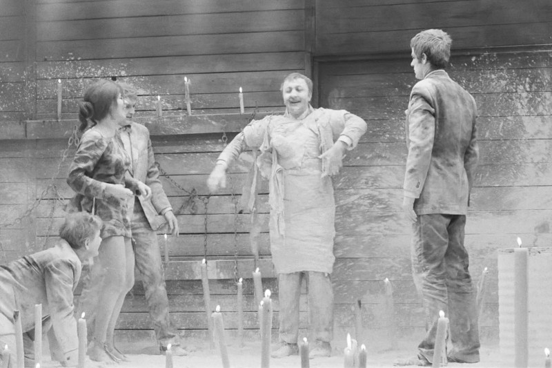 Cztery osoby pokryte kurzem stoją i kucają w kręgu wśród zapalonych świec przed drewnianą ścianą, wydając się odgrywać scenę lub występ. Jedna osoba jest owinięta w materiał i przyjmuje dramatyczną pozę.