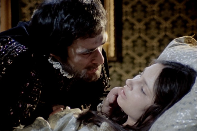 Brodaty mężczyzna w stroju z epoki pochyla się nad młodą kobietą o długich, ciemnych włosach, która leży z zamkniętymi oczami w słabo oświetlonym, ozdobnym pokoju.