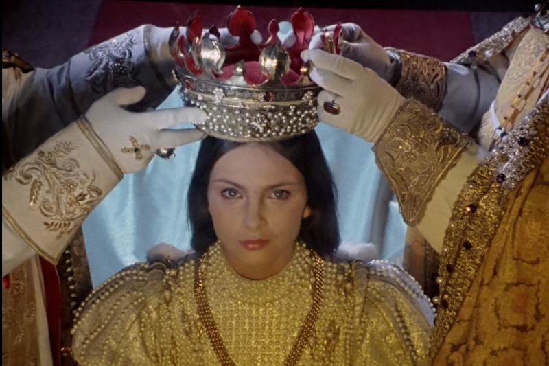 Młoda kobieta w ozdobnych, pokrytych perłami szatach jest koronowana przez dwóch sług w rękawiczkach, trzymających nad głową wysadzaną klejnotami koronę. Spogląda prosto przed siebie ze spokojnym, uroczystym wyrazem twarzy.