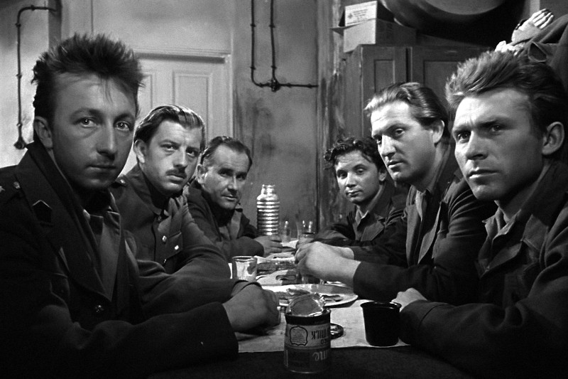 Sześciu mężczyzn w mundurach wojskowych siedzi blisko siebie przy stole, patrząc w stronę kamery z poważnymi minami. Na stole stoi puszka, kubki i czajnik. Sceneria wydaje się być skromnym, industrialnym pokojem.