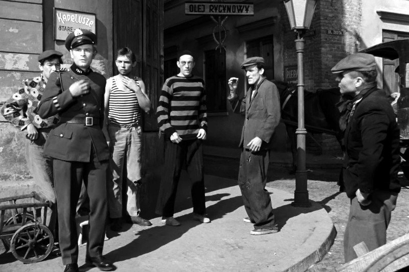 Umundurowany oficer stoi z czterema swobodnie ubranymi mężczyznami na brukowanej ulicy w pobliżu narożnego budynku; grupa wydaje się być zaangażowana w napiętą rozmowę. W tle widać znaki i latarnię uliczną.