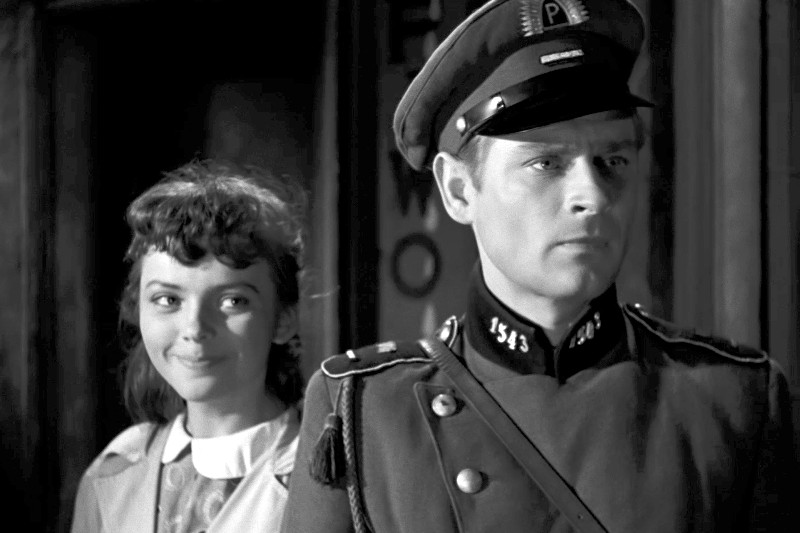 Młoda kobieta uśmiecha się, stojąc za poważnie wyglądającym policjantem w mundurze, na czarno-białej scenie, prawdopodobnie z klasycznego filmu lub programu telewizyjnego.