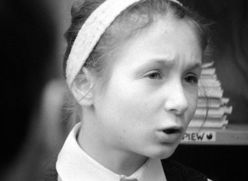 Czarno-białe zdjęcie młodej dziewczyny z opaską na głowie, która wydaje się mówić lub śpiewać, ze skupionym wyrazem twarzy. Na rozmytym tle widać stosy książek.