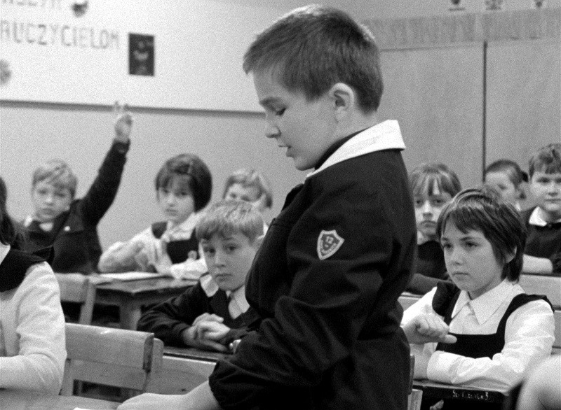 Czarno-białe zdjęcie dzieci w klasie. Jeden uczeń stoi przy ławce i mówi, podczas gdy inni siedzą przy swoich ławkach. Kilku uczniów słucha uważnie, a jeden ma podniesioną rękę.