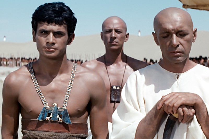 Trzech mężczyzn w starożytnych egipskich strojach stoi na pustyni; mężczyzna po lewej jest z gołą koszulą i nosi ozdobny kołnierz, podczas gdy dwaj pozostali, obaj łysi, mają na sobie białe szaty i wyglądają poważnie.