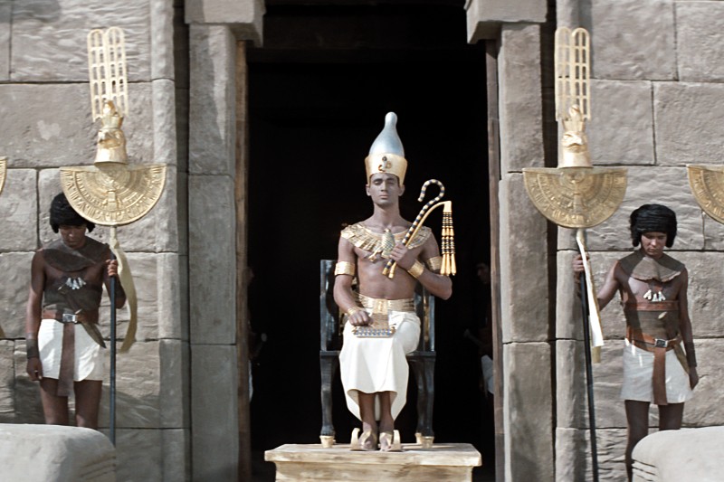 Osoba ubrana jak starożytny egipski faraon siedzi na tronie przed kamienną świątynią, trzymając królewskie berła. Po obu stronach stoją dwaj strażnicy, ubrani w białe spódnice i nakrycia głowy, trzymający wysokie, ozdobne złote laski.