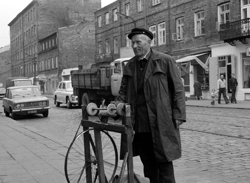 Mężczyzna w płaszczu i czapce stoi przy kole rowerowym i narzędziu do ostrzenia na brukowanej ulicy ze starszymi samochodami, ciężarówką i ceglanymi budynkami w tle. Czarno-białe zdjęcie, prawdopodobnie z połowy XX wieku.