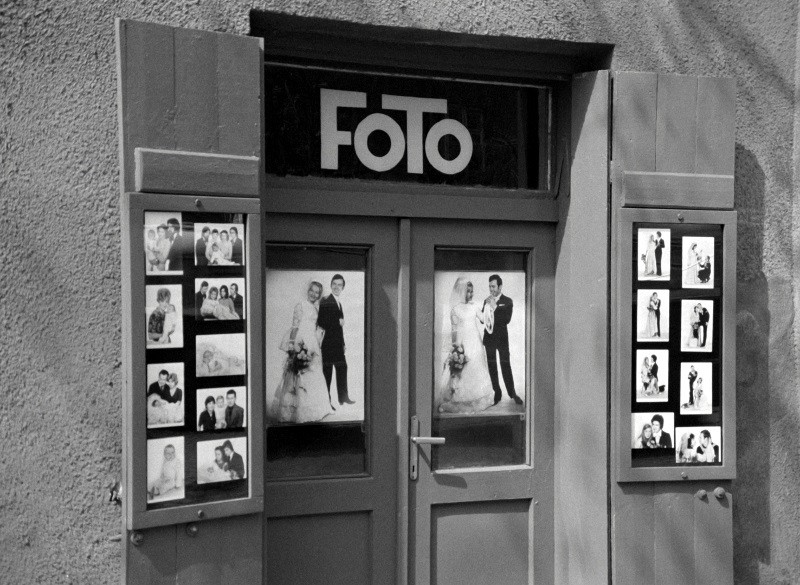 Czarno-białe zdjęcie wejścia do studia fotograficznego z napisem FOTO nad drzwiami oraz oprawionymi w ramki zdjęciami ślubnymi i portretowymi par i rodzin na drzwiach i okiennicach.