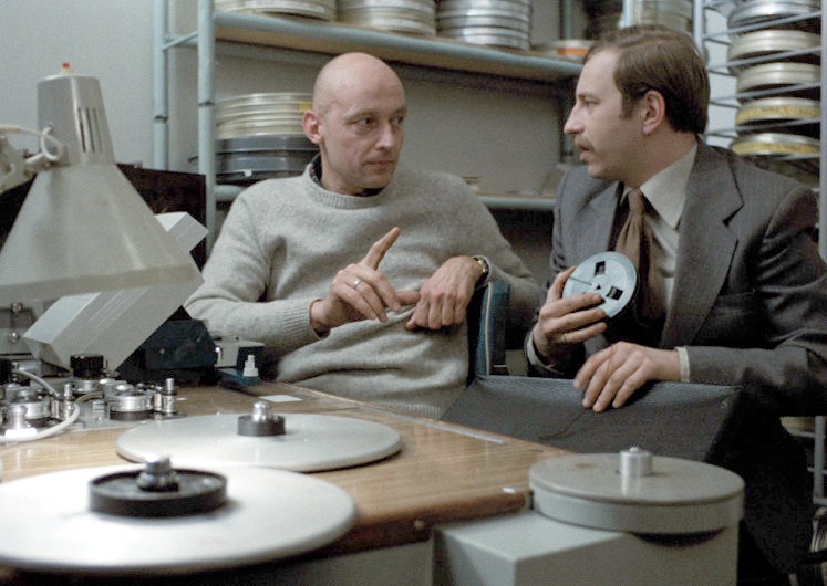 Dwóch mężczyzn siedzi w pokoju montażowym, otoczonych szpulami i sprzętem. Jeden z nich gestykuluje ręką, podczas gdy drugi trzyma szpulę, a oni wydają się być w rozmowie, skupieni i zaangażowani.