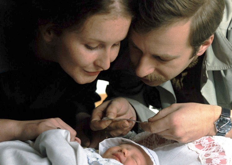 Kobieta i mężczyzna delikatnie pochylają się nad noworodkiem owiniętym w kocyk, patrząc na niemowlę z uczuciem. Mężczyzna trzyma przedmiot w pobliżu dziecka, a oboje dorośli delikatnie się uśmiechają.