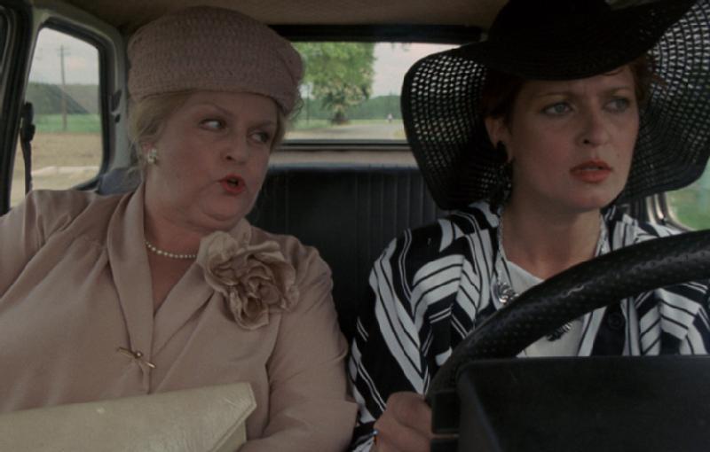 Dwie kobiety siedzą na przednich siedzeniach samochodu. Pasażerka ubrana w jasny strój i kapelusz rozmawia, podczas gdy kierowca, ubrany w czarny kapelusz i pasiastą kurtkę, patrzy przed siebie z poważnym wyrazem twarzy.