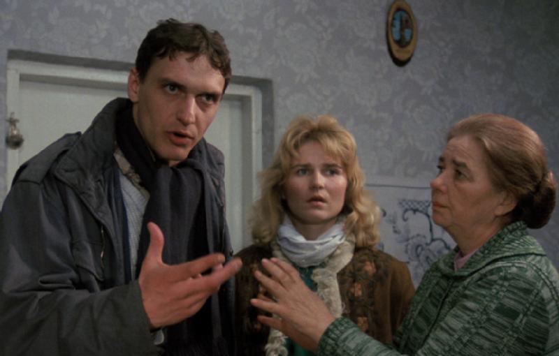 Poważnie wyglądający mężczyzna gestykuluje, stojąc obok zaniepokojonej młodej kobiety i starszej kobiety. Wszystko to znajduje się w środku, na tle wzorzystej tapety.