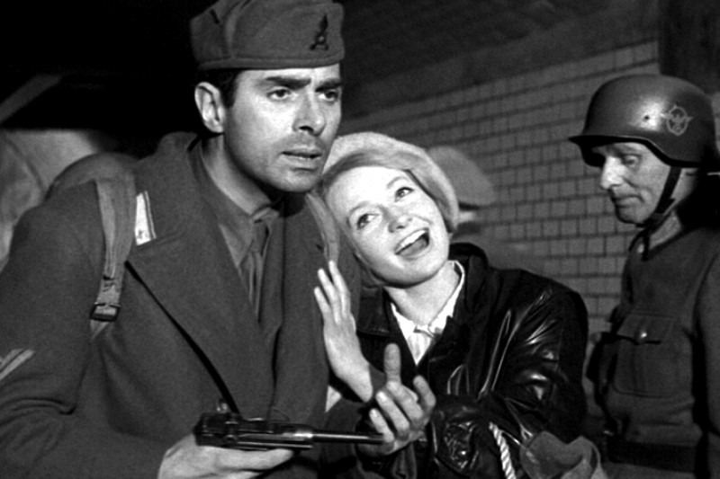 Mężczyzna w mundurze wojskowym trzymający pistolet stoi obok uśmiechniętej kobiety, która opiera głowę na jego ramieniu; w tle stoi inny żołnierz w hełmie. Scena jest czarno-biała.