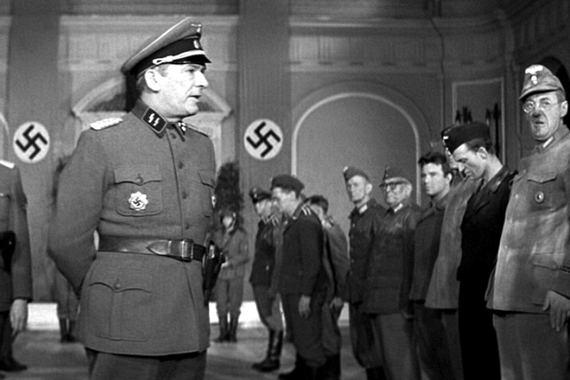 Grupa mężczyzn w mundurach wojskowych z czasów II wojny światowej stoi w dużej hali, na której ścianach wiszą transparenty ze swastykami. Na pierwszym planie stoi umundurowany oficer, zwracając się do pozostałych osób ustawionych za nim.