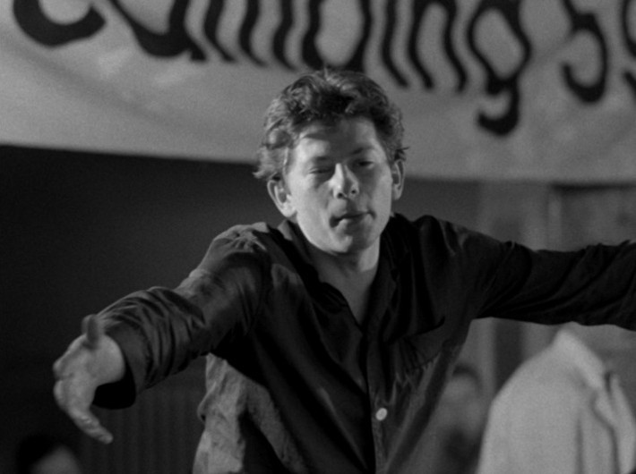Młody mężczyzna z krótkimi włosami i ciemną koszulą stoi z wyciągniętymi ramionami, sprawiając wrażenie, jakby był w ruchu. W tle wisi częściowo widoczny baner z dużym napisem. Obraz jest czarno-biały.