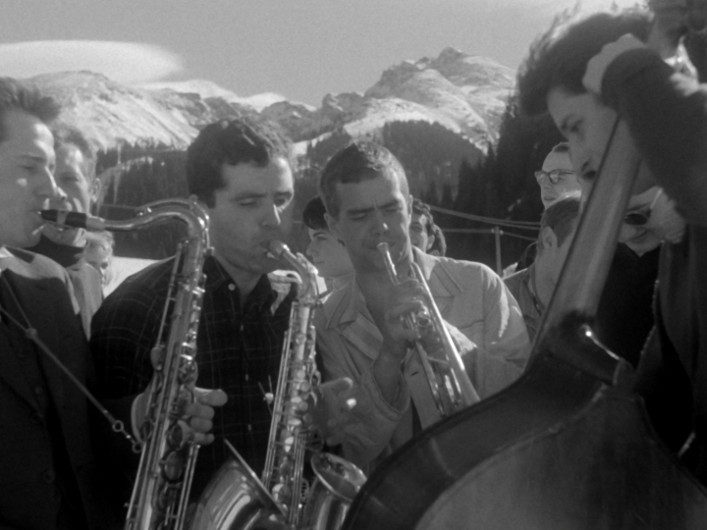 Grupa młodych mężczyzn gra na saksofonach, trąbce i kontrabasie na świeżym powietrzu w górach, w tle widać ośnieżone szczyty.