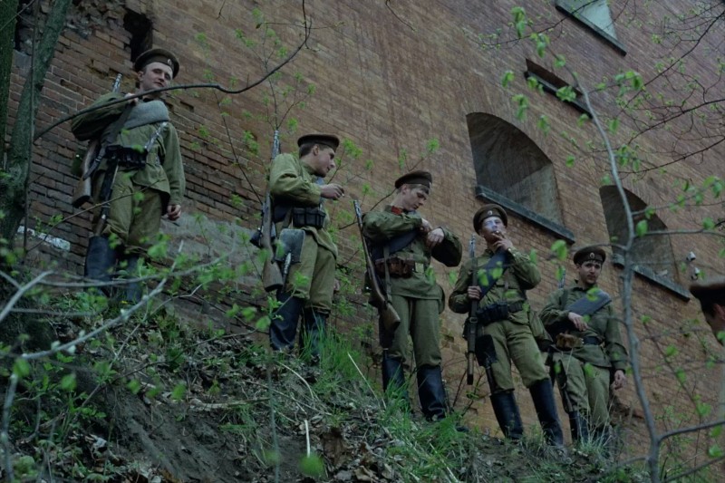 Pięciu żołnierzy w zabytkowych mundurach wojskowych stoi na trawiastym zboczu w pobliżu ceglanego budynku, trzymając karabiny i patrząc w dół. Scena wydaje się rozgrywać podczas minionej wojny, z drzewami i liśćmi na pierwszym planie.