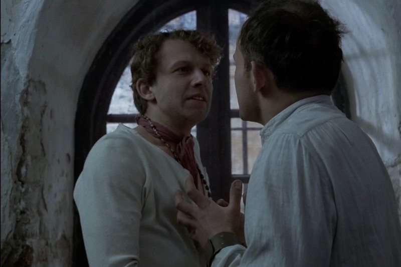 Dwóch mężczyzn stoi naprzeciwko siebie przed łukowatym oknem, pogrążonych w napiętej rozmowie. Jeden z nich dotyka piersi drugiego, a obaj mają na sobie staromodne, jasne koszule. W tle widać stare, łuszczące się ściany.