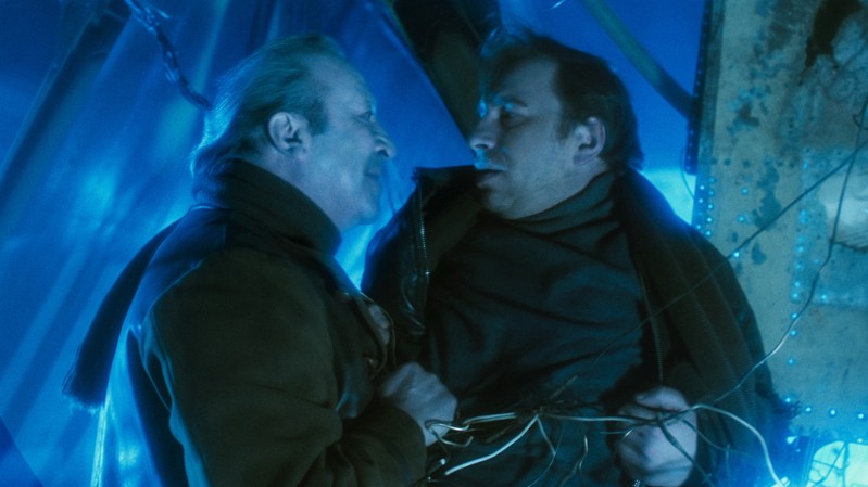 Dwóch mężczyzn staje naprzeciw siebie w intensywnym niebieskim świetle; jeden chwyta drugiego za płaszcz i unosi go nad ziemię, przygważdżając drutami do metalowej konstrukcji.