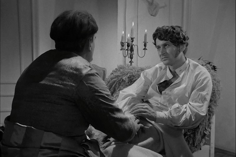 Dwóch mężczyzn w strojach z epoki siedzi w słabo oświetlonym pokoju. Jeden z nich stoi twarzą do kamery, odchyla się na krześle i zamyślony patrzy na drugiego mężczyznę, który siedzi tyłem do widza. W tle widać świecznik.