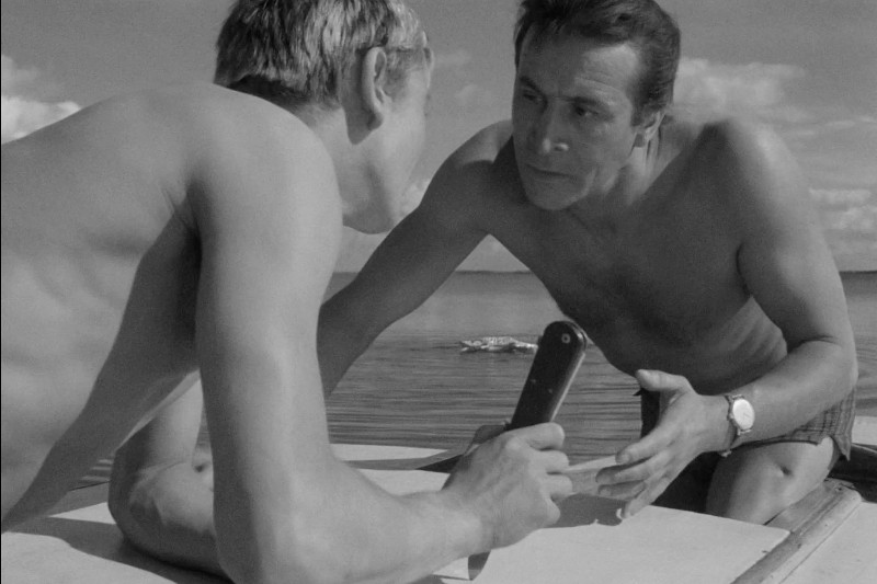 Dwóch mężczyzn bez koszuli na łodzi prowadzi intensywną rozmowę; jeden trzyma składany nóż, pochylając się w stronę drugiego. Scena rozgrywa się na spokojnej wodzie pod częściowo zachmurzonym niebem.