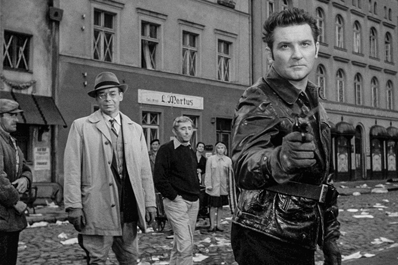 Mężczyzna w skórzanej kurtce celuje bronią w kamerę na zaśmieconej ulicy, podczas gdy kilka osób stoi za nim, patrząc z napiętymi minami. W tle widać stare budynki z wybitymi oknami.