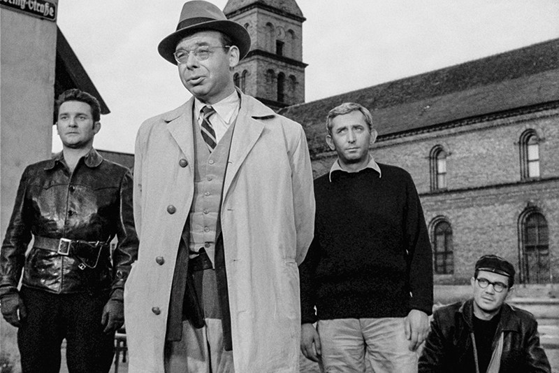 Czterech mężczyzn stoi na zewnątrz przed dużym kamiennym budynkiem. Mężczyzna z przodu nosi trencz, krawat i kapelusz. Trzech mężczyzn za nim nosi ciemniejsze, bardziej swobodne ubrania i patrzy w różnych kierunkach. Obraz jest czarno-biały.