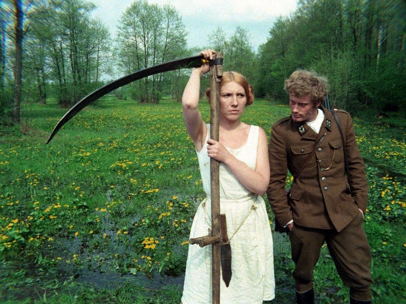 Kobieta w białej sukience trzyma dużą kosę na trawiastym, pełnym kwiatów polu. Obok niej mężczyzna w brązowym mundurze opiera się o kosę, patrząc na nią. Wysokie drzewa są w tle pod zachmurzonym niebem.