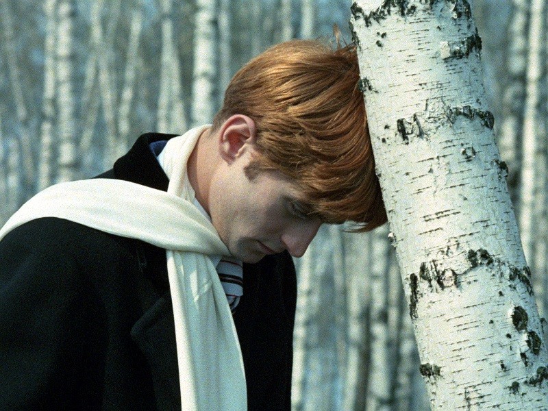 Osoba o rudych włosach, ubrana w ciemny płaszcz i biały szalik, opiera czoło o białą brzozę w lesie podobnych drzew, sprawiając wrażenie zamyślonej lub ponurej.