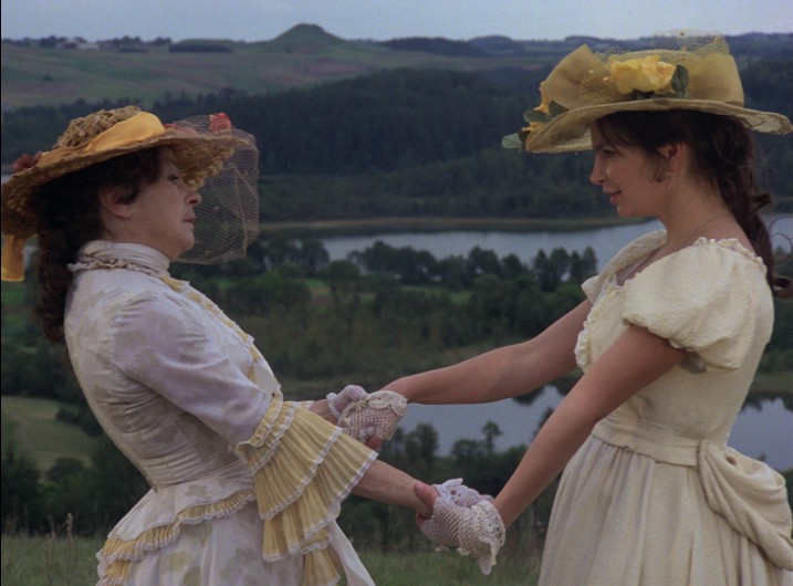 Dwie kobiety w kremowych sukienkach vintage i słomkowych kapeluszach, trzymające się za ręce i zwrócone ku sobie, stojące na świeżym powietrzu, na trawiastym wzgórzu, z malowniczym krajobrazem drzew, wody i odległych wzgórz w tle.