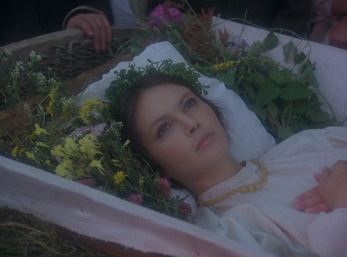 Kobieta z wiankiem z kwiatów i naszyjnikiem leży w trumnie otoczonej kwiatami, patrząc w górę z pogodnym wyrazem twarzy. Jej ręce są złożone na piersi, a ona sama jest ubrana na biało.