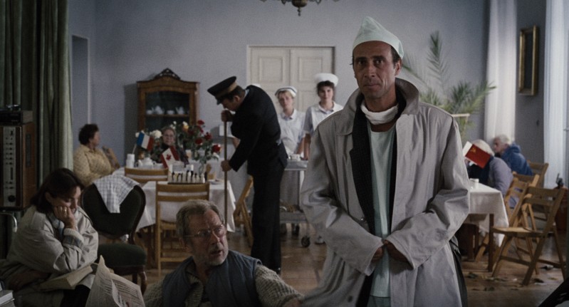 Mężczyzna w białym fartuchu i szpitalnym czepku stoi w pokoju z pacjentami siedzącymi przy stołach. Pielęgniarki i mężczyzna w mundurze są w tle. Atmosfera jest ponura, z przytłumionymi kolorami i stonowanym oświetleniem.