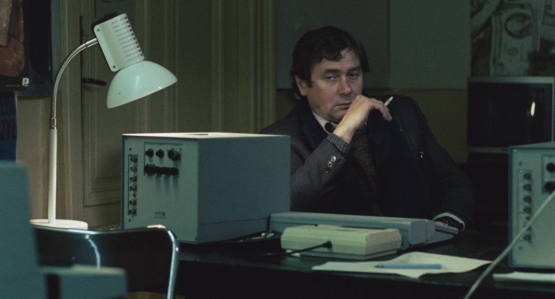 Mężczyzna w garniturze siedzi przy biurku w słabo oświetlonym biurze, opierając brodę na dłoni i trzymając papierosa. Otacza go zabytkowy sprzęt elektroniczny i biała lampa biurkowa.