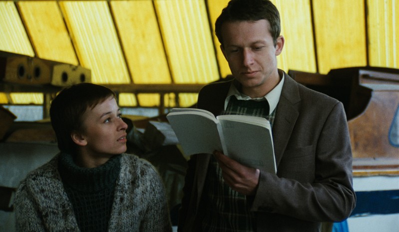 Kobieta z krótkimi włosami spogląda na mężczyznę w garniturze, który czyta z otwartej książki. Są w środku, z przyciemnianymi na żółto oknami i metalowymi konstrukcjami w tle.